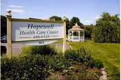 Outdoor view of Hopewell Health Care Center sign with phone number and address, surrounded by greenery and bushes, with a white gazebo and trees in the background under a clear sky.