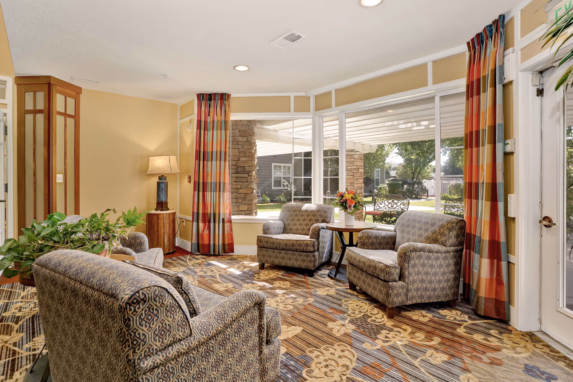 A cozy living room area with patterned armchairs and a sofa arranged around a small wooden table with a flower vase. Large windows with colorful striped curtains let in natural light and offer a view of an outdoor garden with benches and trees. A floor lamp and a potted plant add to the warm and inviting atmosphere.