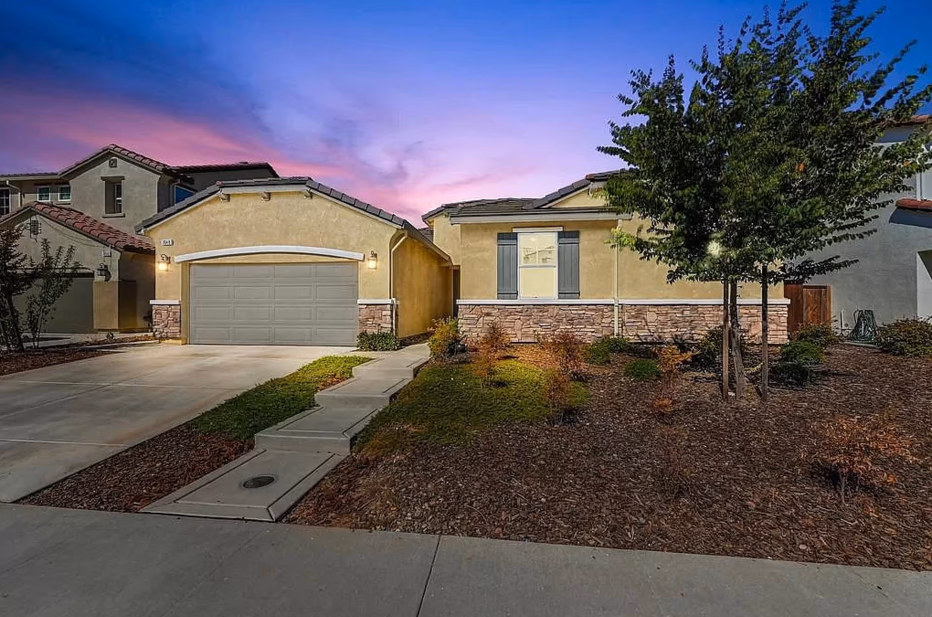 Exterior view of a single-story residential building at dusk with a two-car garage, a concrete driveway, a small front yard with mulch and young trees, and a sidewalk leading to the front door. The sky is a gradient of blue and purple colors.