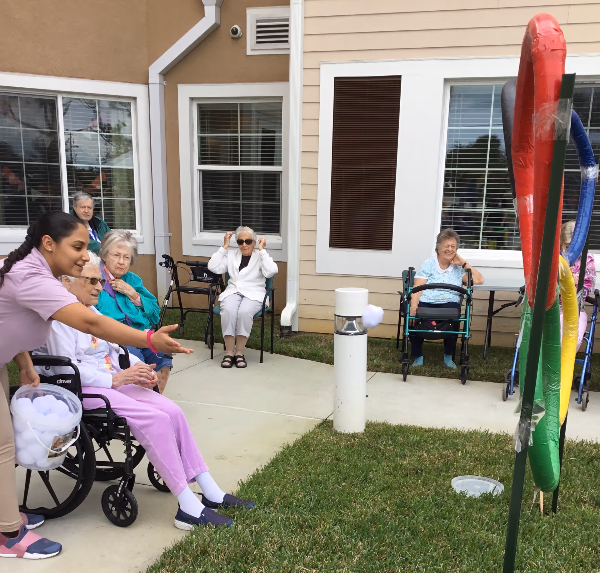 A group of elderly women, some seated in wheelchairs and others in chairs, are outdoors at an assisted living facility. A caregiver is helping one of the women throw soft balls through colorful rings set up on a lawn. The building with windows and beige siding is visible in the background.
