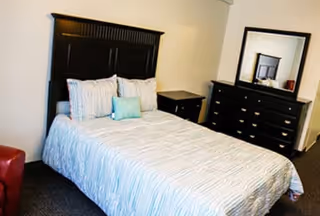A bedroom with a neatly made bed featuring a striped comforter and multiple pillows. The bed has a dark wooden headboard. Next to the bed is a matching dark wooden nightstand. Across from the bed is a dark wooden dresser with multiple drawers and a large mirror on top. The room has neutral-colored walls and carpeted flooring.