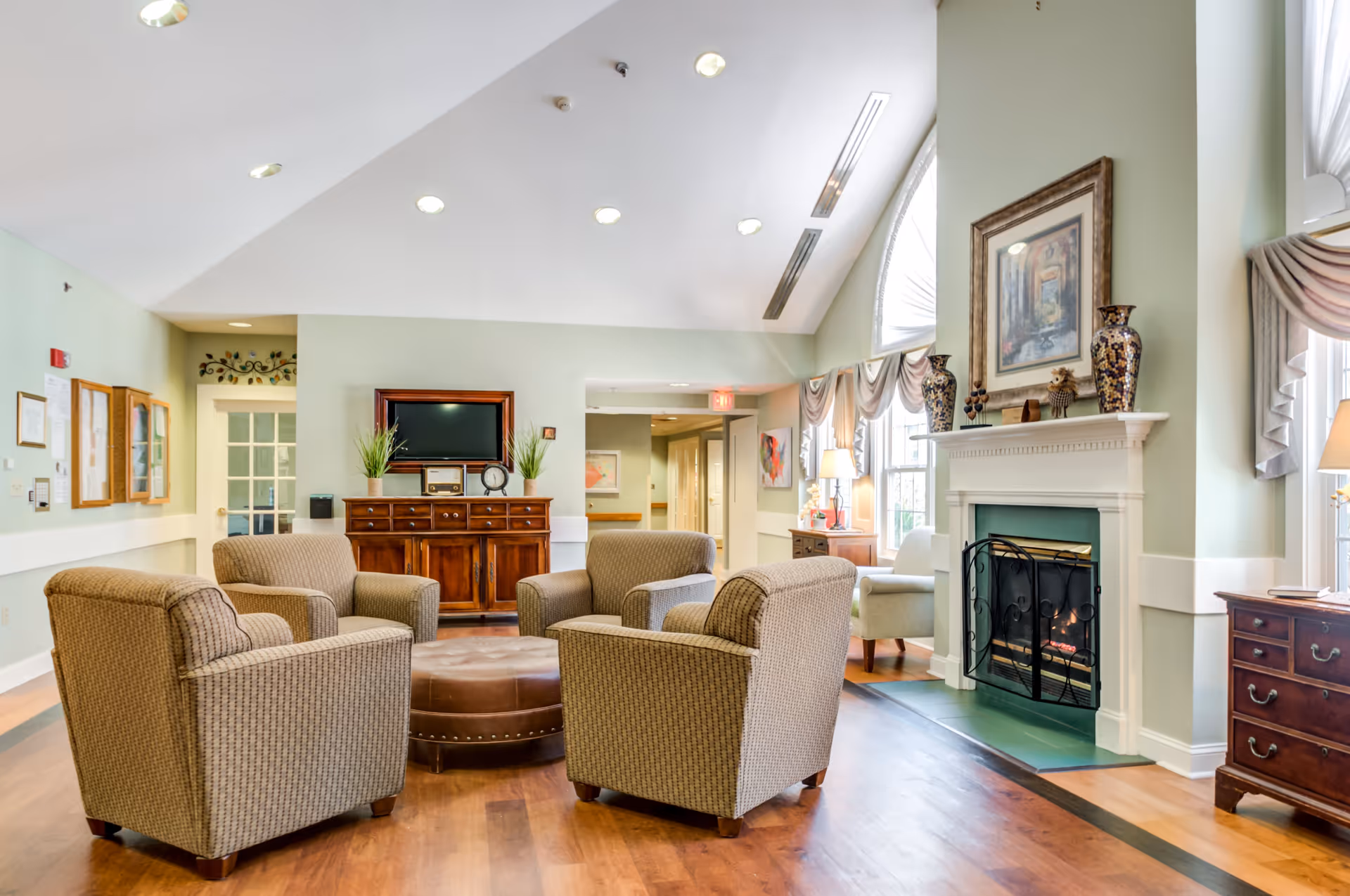 A cozy living room area with four upholstered armchairs arranged around a round leather ottoman. The room features a fireplace with decorative vases and a framed painting above it, large windows with draped curtains, a wooden cabinet with a TV and plants, and hardwood flooring under a high vaulted ceiling with recessed lighting.