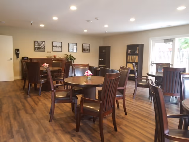 A well-lit dining area with several round wooden tables and matching chairs arranged neatly on a wood floor. Each table has a small floral centerpiece. The room has beige walls with framed black and white photos, a bookshelf, and a door leading outside.