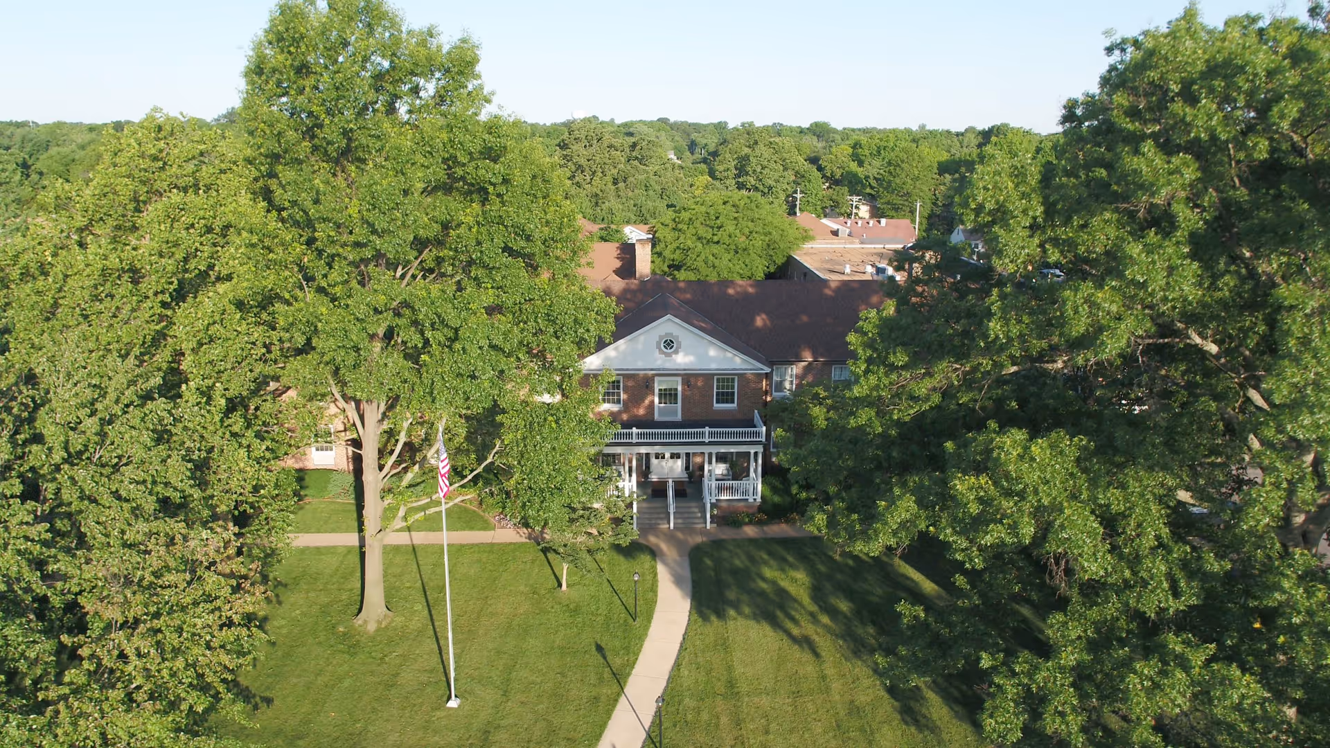 Aerial view of a senior living facility named Manor Grove showing a large brick building with a white porch entrance surrounded by green trees and a well-maintained lawn with a pathway leading to the entrance. An American flag is visible on a flagpole in front of the building.