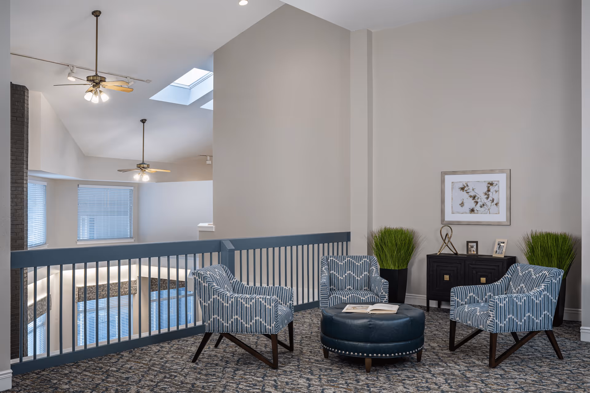 A cozy seating area in a senior living facility with three blue patterned armchairs arranged around a round blue ottoman with an open book on it. The space features a blue railing overlooking a lower level, two ceiling fans with lights, skylights, and neutral-colored walls. There is a black cabinet with decorative items and two tall green plants on either side, along with a framed artwork on the wall.