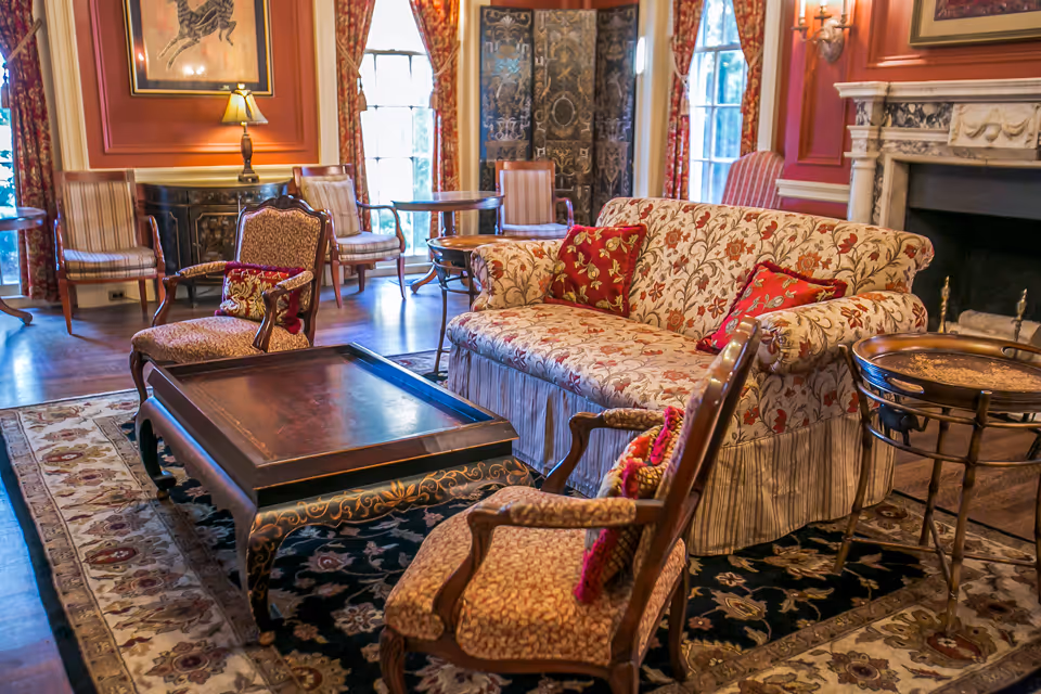 A cozy living room with floral-patterned sofa and matching armchairs, a dark wooden coffee table, ornate area rugs, a fireplace with a marble mantel, and large windows with red patterned curtains letting in natural light.