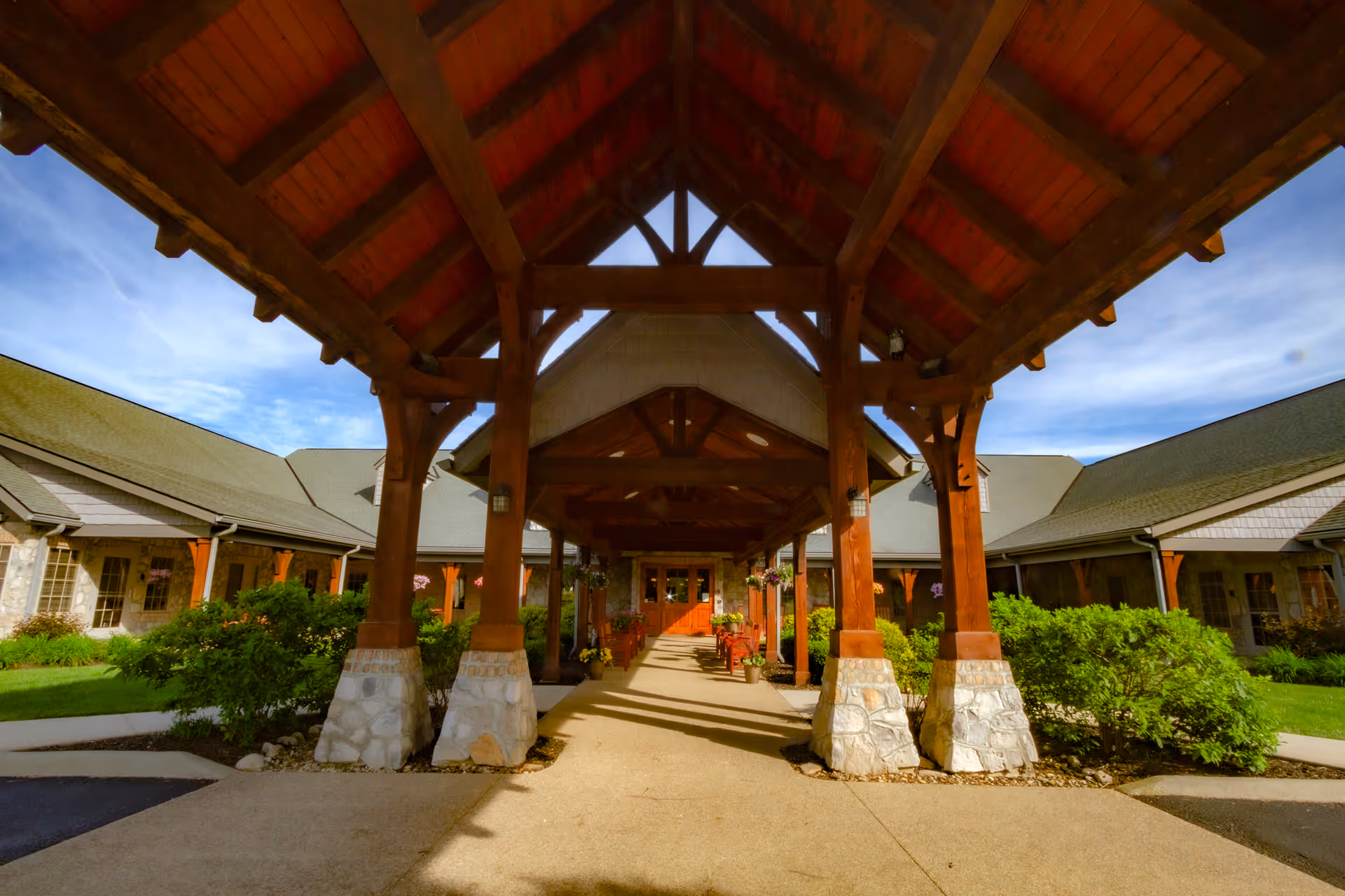 Covered entrance walkway with wooden beams and stone bases leading to the main building of the Inn at Bear Trail, surrounded by green bushes and a clear blue sky.