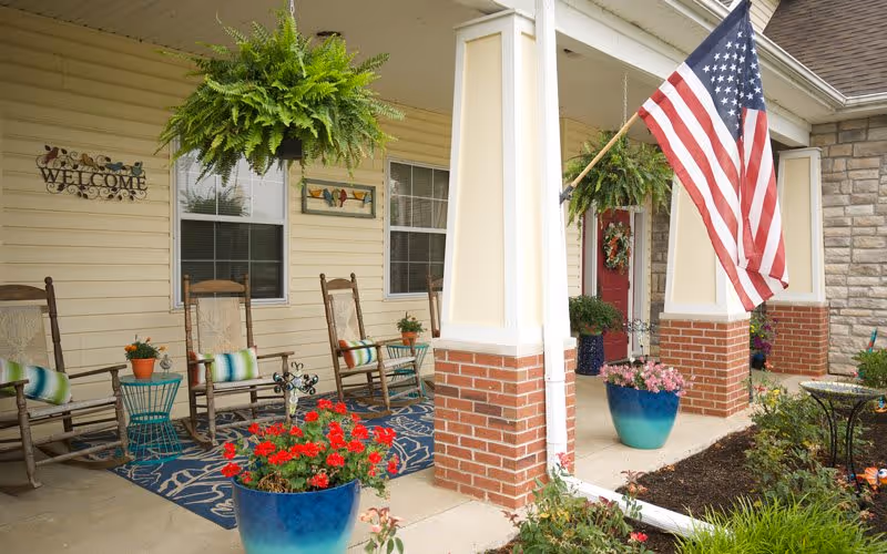 Front porch of a building with beige siding and brick pillars, decorated with hanging green ferns, an American flag, rocking chairs with cushions, potted plants with red and pink flowers, and a welcome sign on the wall near two windows.