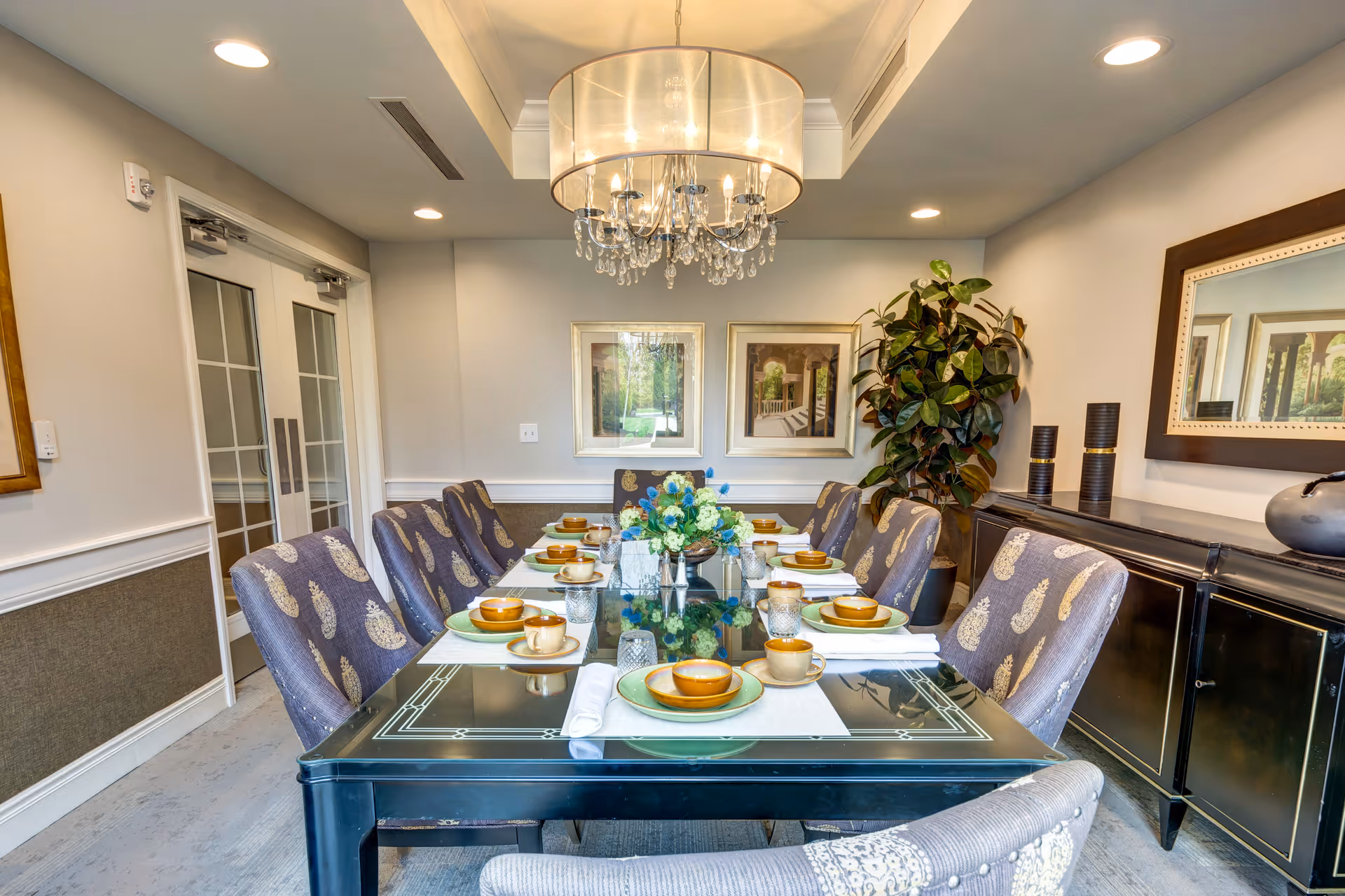 A formal dining room with a long black glass table set for eight people. Each place setting includes a green plate, a brown bowl, a brown cup, and a white napkin. The chairs are upholstered in a patterned fabric. A chandelier with crystal accents hangs above the table. The room has light-colored walls with two framed pictures and a large mirror. There is a large potted plant in the corner and a black sideboard with decorative items.