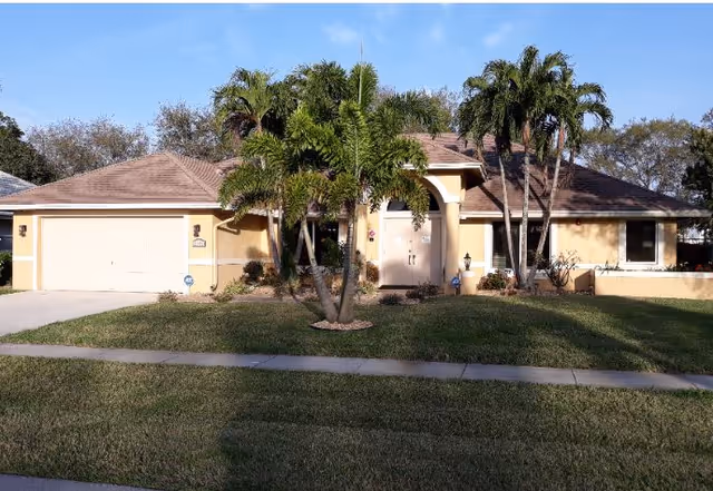 Single-story beige house with a two-car garage, a front door with an archway, and several palm trees in the front yard. The lawn is green and there is a sidewalk in front of the house.