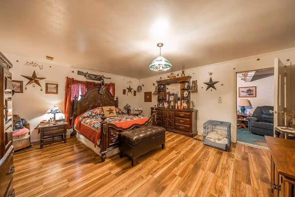 A warmly decorated bedroom with wooden flooring, a large wooden bed with a red and patterned quilt, a matching wooden dresser with a mirror, and a nightstand with a lamp. The walls are adorned with star-shaped decorations and framed pictures. There is a dog crate with a cushion inside near the open door, which leads to a living room area with a blue couch and a lamp on a side table.