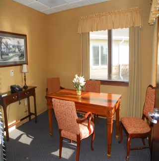 A small room with a wooden table and four upholstered chairs. There is a window with beige curtains letting in natural light. A small wooden side table with a lamp and a framed picture on the wall are also visible.