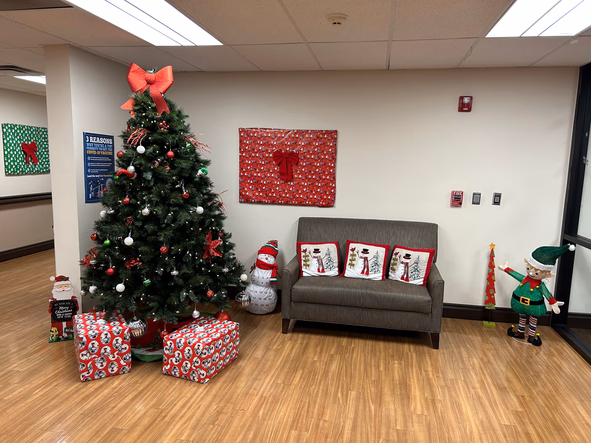 A festive holiday corner in a healthcare facility featuring a decorated Christmas tree with red, white, and green ornaments and a large red bow on top. There are wrapped presents under the tree, a gray loveseat with three snowman-themed pillows, and holiday decorations including a snowman figure, a small Christmas tree decoration, and an elf figure. The floor is wood, and the walls are plain with a few holiday-themed wall hangings.