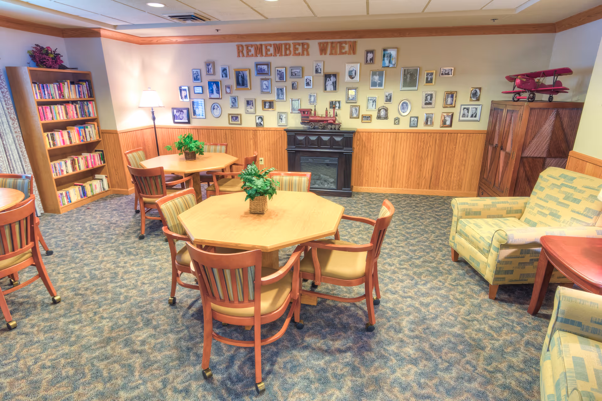 A cozy senior living common area with carpeted floor, wooden tables and chairs, a bookshelf filled with books, a wall decorated with numerous framed photos and the words 'REMEMBER WHEN' above them, a floor lamp, two patterned armchairs, and a small fireplace with a decorative red airplane model on a cabinet nearby.