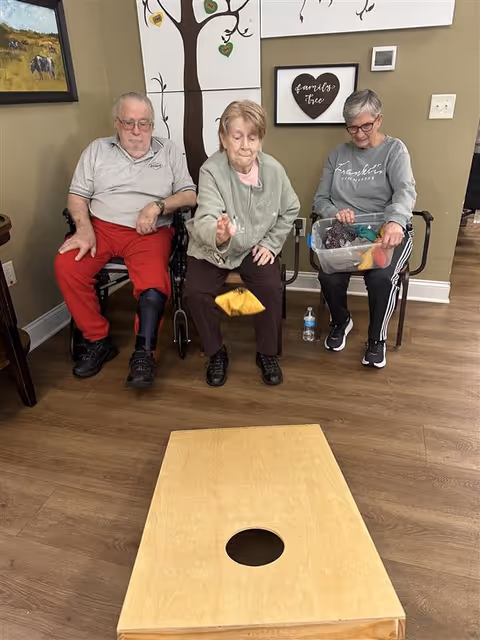 Three elderly individuals sitting in chairs indoors playing a game of cornhole. The person in the middle is tossing a bean bag towards a wooden cornhole board with a hole. The room has wooden flooring and wall decorations including a tree painting and a framed sign that says 'family tree'.