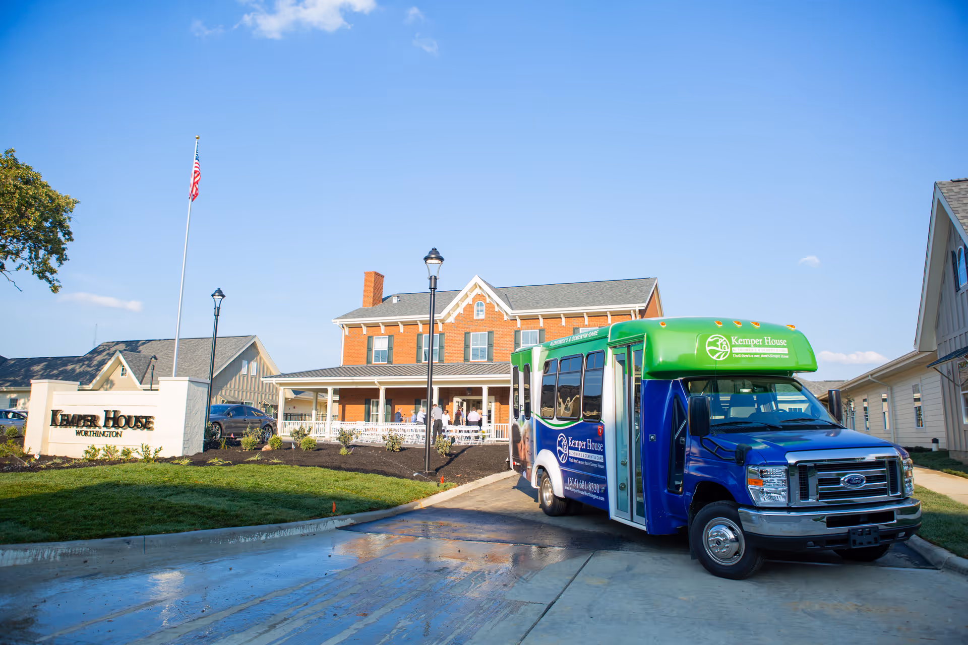 Exterior view of Cotter House Worthington, a senior living facility, showing a brick building with a porch, an American flag on a flagpole, and a blue and green Kemper House shuttle bus parked in front.
