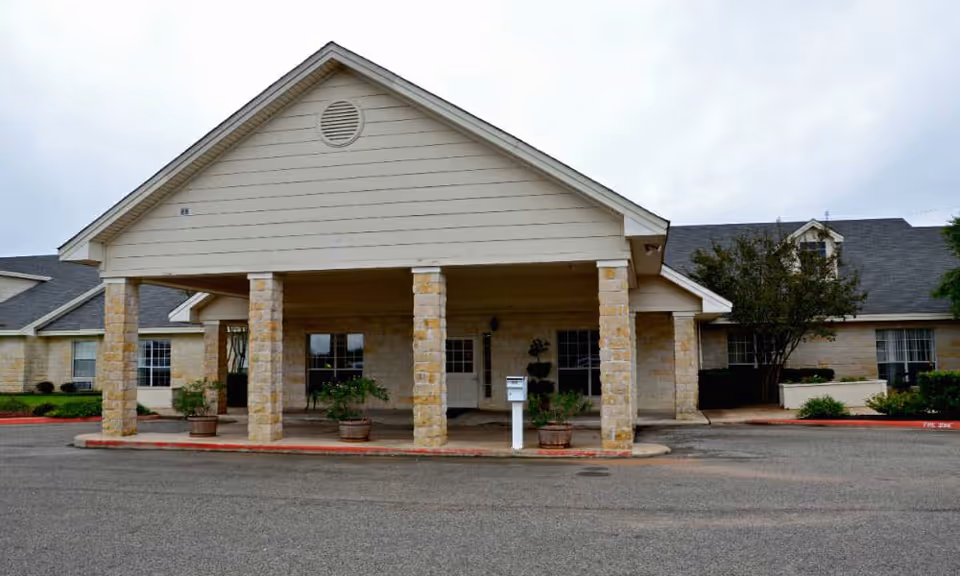 Front exterior view of a single-story building with a covered entrance supported by stone pillars, surrounded by a paved driveway and some landscaping with bushes and small trees.
