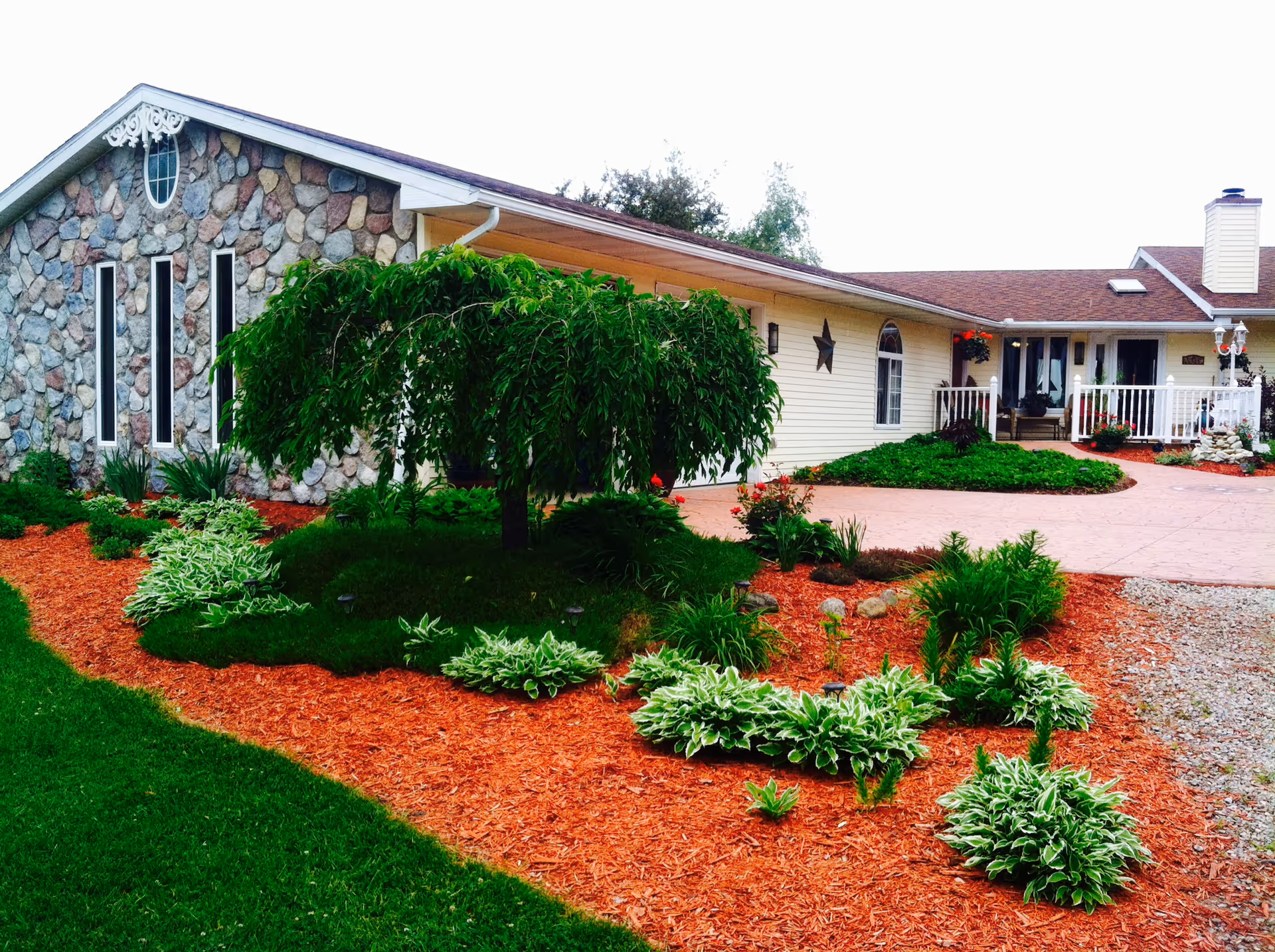 Exterior view of a single-story building with a stone facade on one side and beige siding on the other. The building has a porch with white railings and a chimney. In front, there is a landscaped garden with a variety of green plants, a small tree, and red mulch covering the ground. The driveway is paved and curves around the garden area.