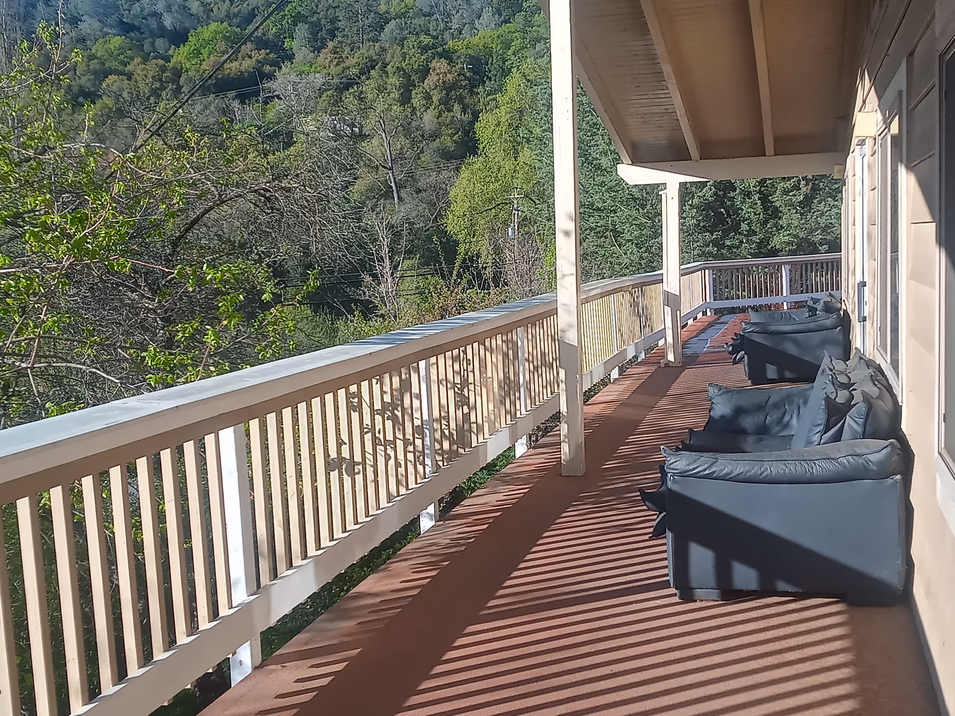 A covered outdoor balcony with wooden railings overlooking a lush green forested area. The balcony has several dark cushioned armchairs arranged along the wall of the building, with sunlight casting shadows through the railings onto the floor.