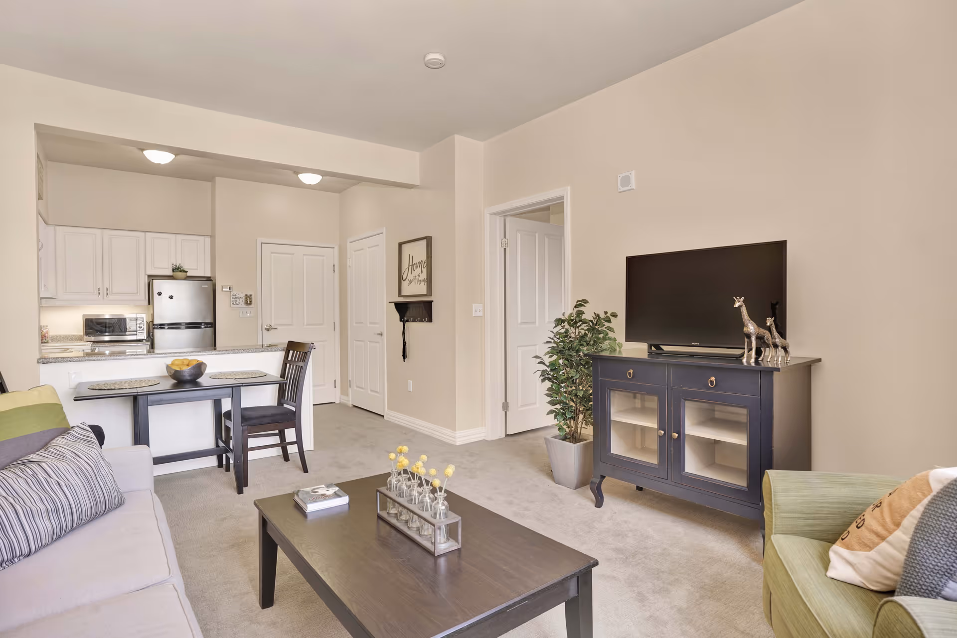 A cozy living room with a beige sofa and green armchair, a dark wooden coffee table with decorative items, a dark wooden TV stand with a flat-screen TV and giraffe figurines, a potted plant, and an open view into a small kitchen area with white cabinets, a stainless steel refrigerator, and a dining table with two chairs.