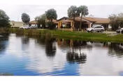 View of a single-story assisted living facility building with a covered entrance, surrounded by palm trees and parked cars, reflected in a calm pond in the foreground.