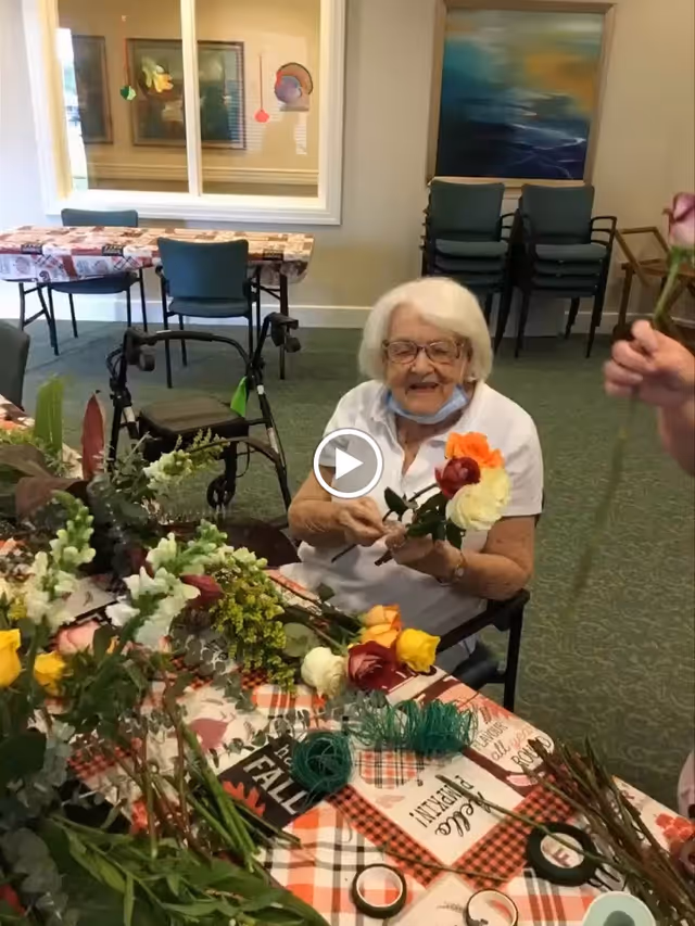 An elderly woman with white hair and glasses sitting at a table covered with a fall-themed tablecloth, arranging colorful flowers. There are various flowers and floral supplies on the table. In the background, there are stacked chairs, a walker, and a window with decorations.