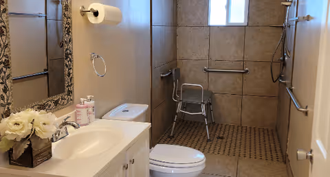 A clean bathroom with beige tiled walls and floor. It features a white sink with a mirror above it, a toilet, a shower area with a shower chair, and multiple grab bars for accessibility. There is a small window in the shower area and a roll of toilet paper on the wall.