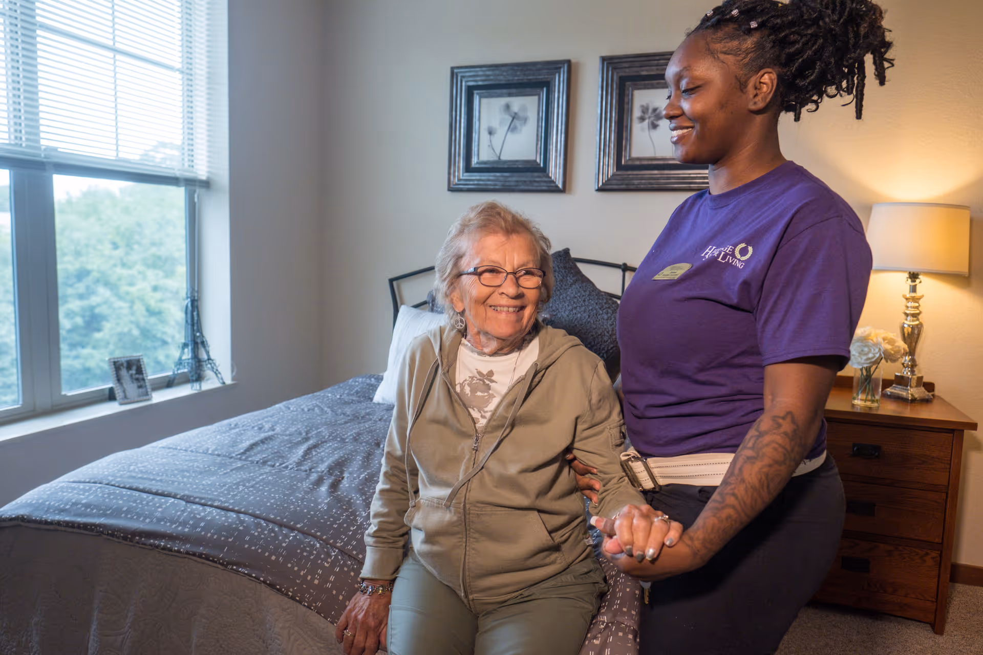 An elderly woman sitting on the edge of a bed in a bedroom, smiling and holding hands with a caregiver standing beside her. The caregiver is wearing a purple shirt with the Heritage Assisted Living logo. The room has a window with blinds, framed artwork on the wall, a bedside table with a lamp and flowers, and a photo frame on the windowsill.