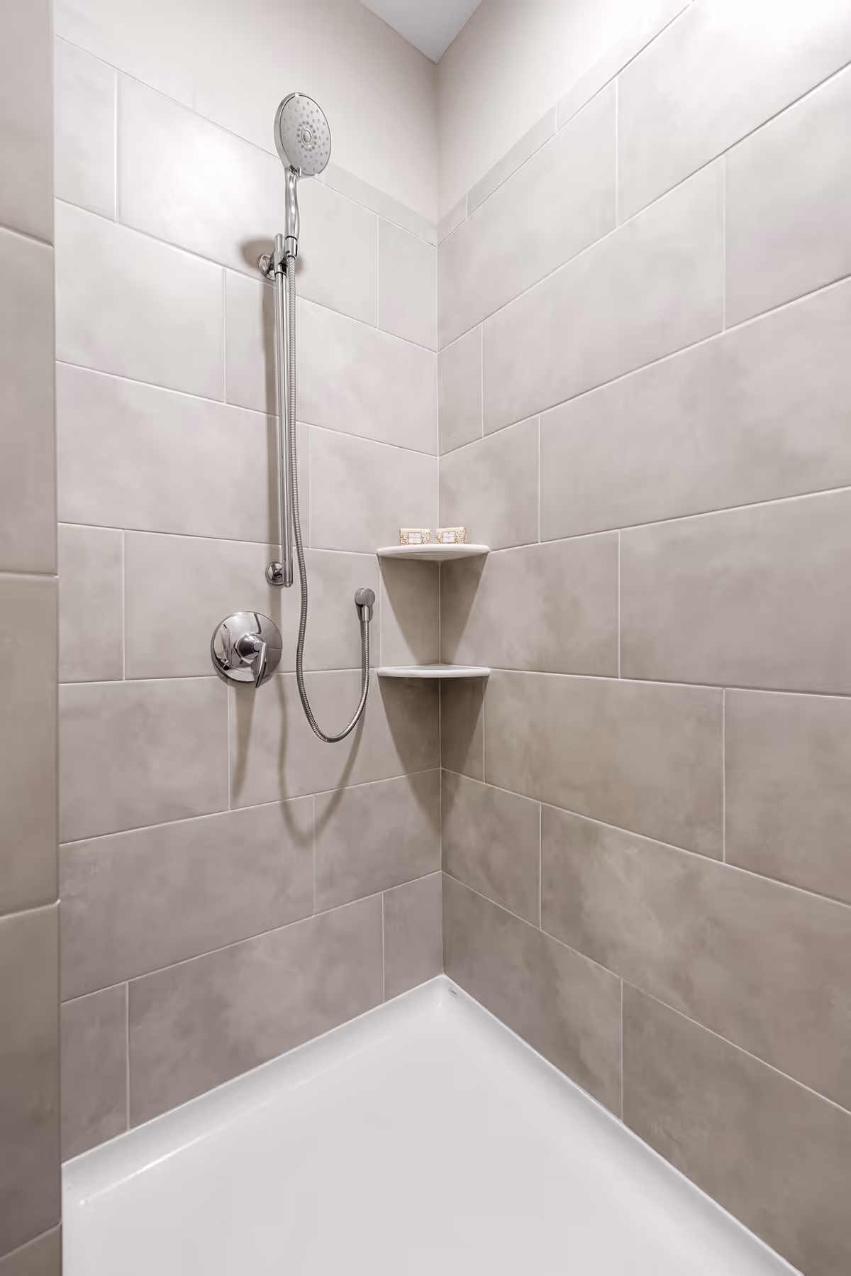 A corner shower with beige tiled walls, a silver handheld showerhead attached to a vertical bar, a control knob, and two small corner shelves holding soap bars.