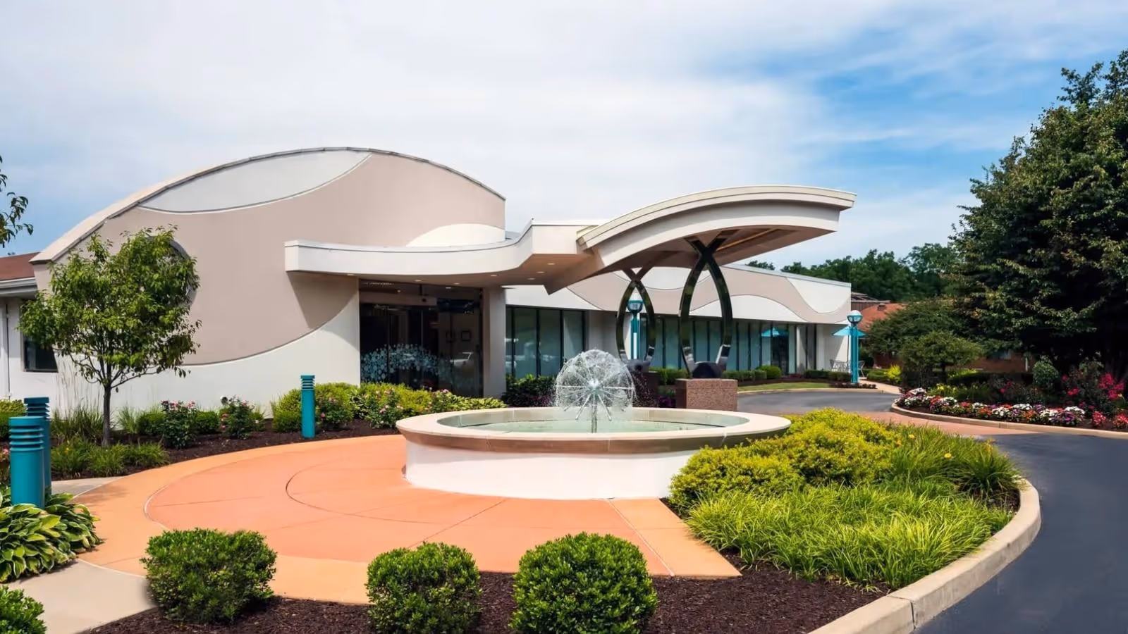 Exterior view of Delmar Gardens On the Green facility featuring a modern building with curved architectural elements, a circular water fountain in the foreground, landscaped greenery, and a curved driveway.