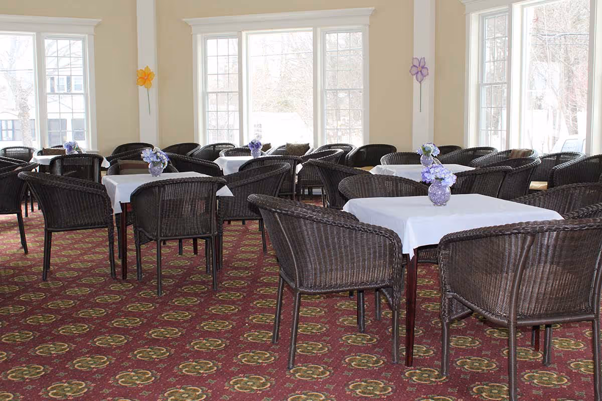 A bright dining room with multiple square tables covered with white tablecloths, each adorned with a small vase of purple flowers. The room has large windows letting in natural light, beige walls, and a patterned red carpet. Dark wicker chairs surround each table.