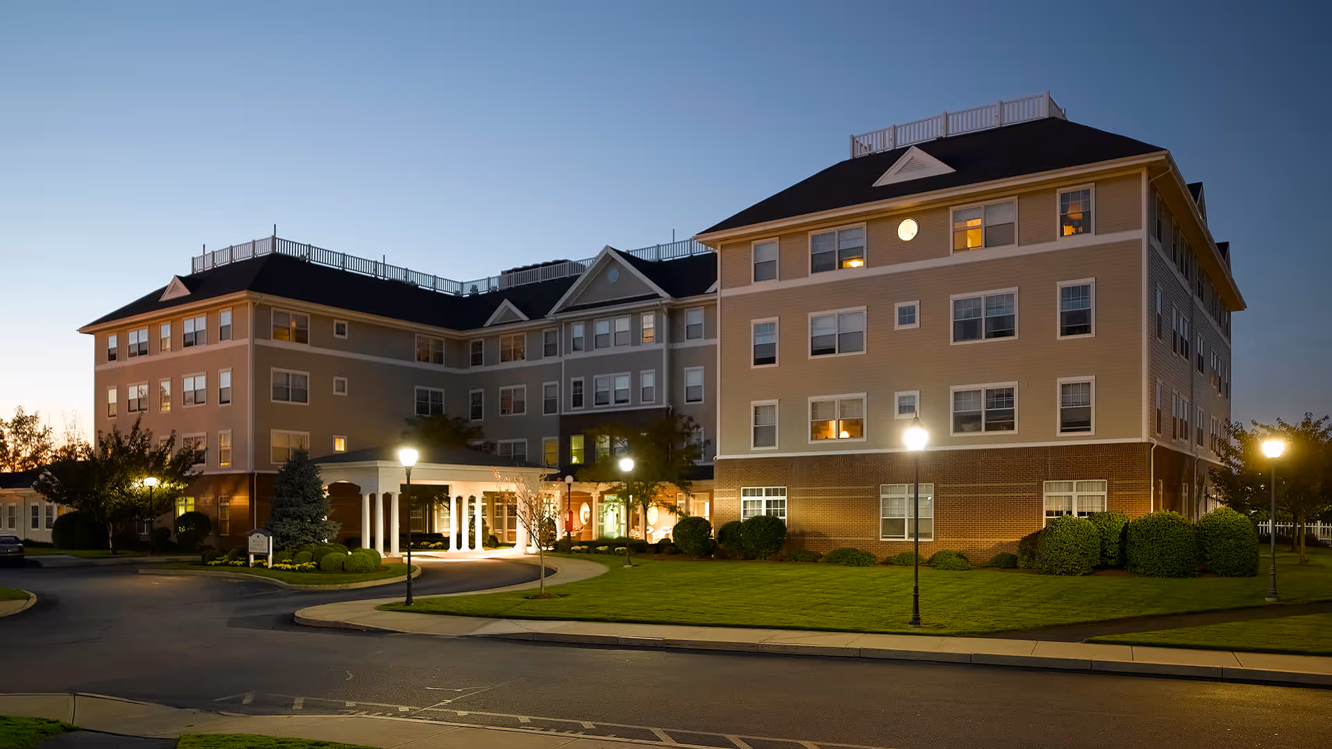 Exterior view of a multi-story senior living facility building named Atria Marina Place at dusk, with lit street lamps, well-maintained lawn, and a covered entrance.