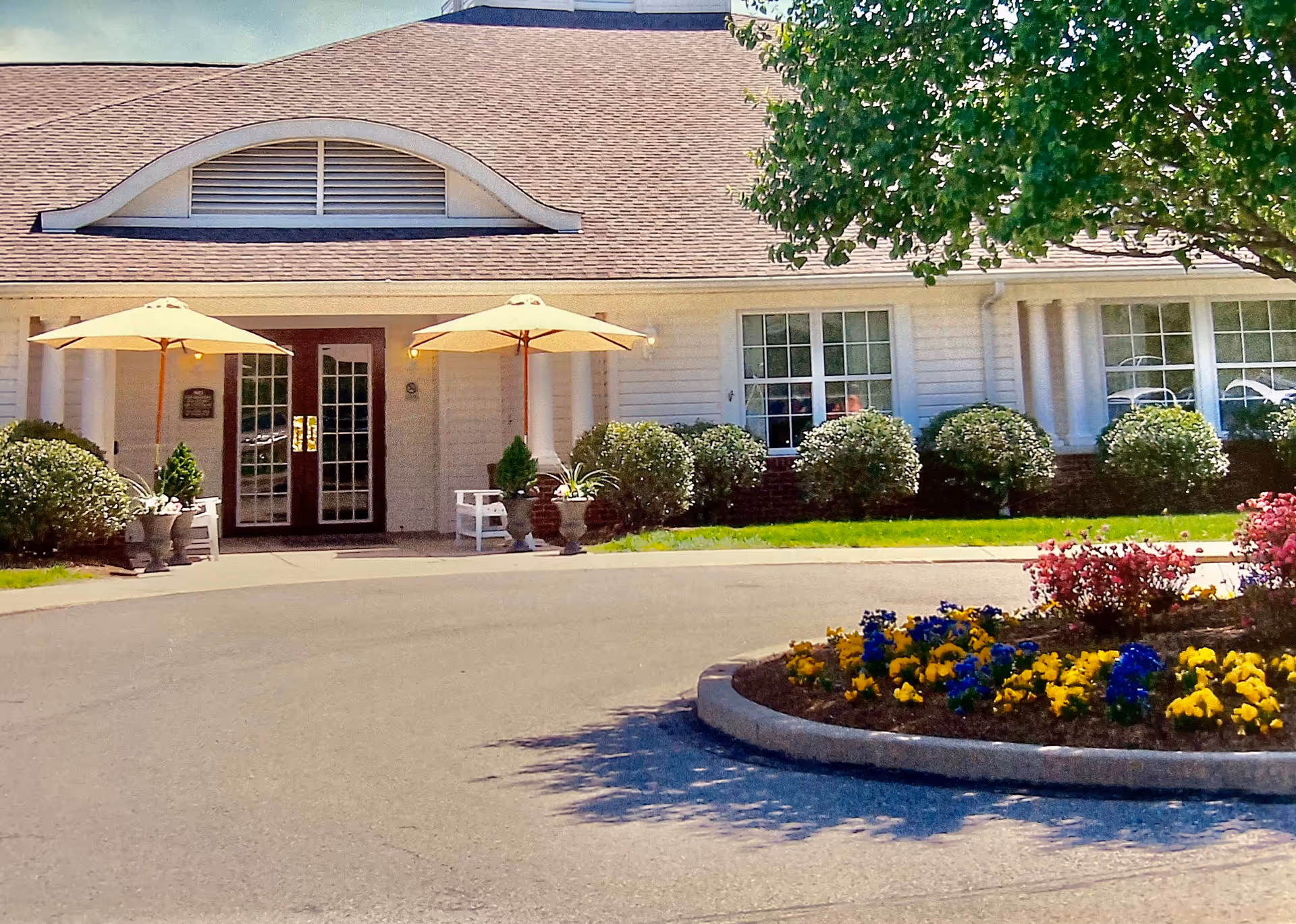 Front exterior view of a single-story building with white siding and a brown shingled roof. The entrance features double glass doors with two large beige umbrellas shading white benches on either side. There are neatly trimmed bushes along the front of the building and a circular flower bed with colorful yellow, blue, and pink flowers in the foreground. A large tree provides shade on the right side of the image.