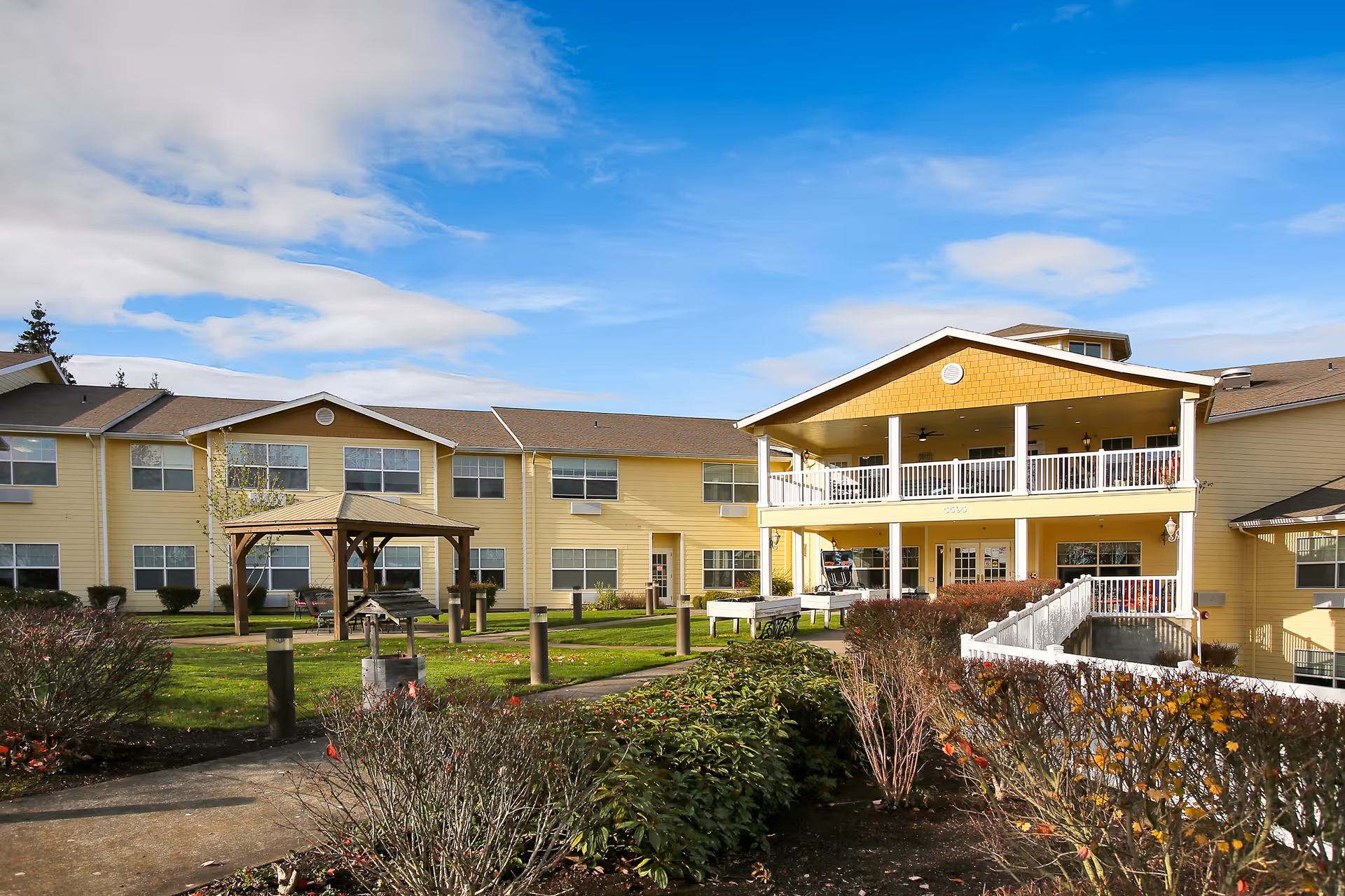 Exterior view of a two-story yellow senior living facility building with multiple windows, a covered porch with white railings, a wooden gazebo, and landscaped bushes and grass under a partly cloudy sky.