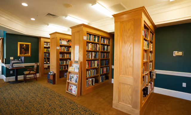 Interior view of a library room in an assisted living facility with wooden bookshelves filled with books, a carpeted floor, green walls with white trim, and a small desk with a chair and computer in the background.