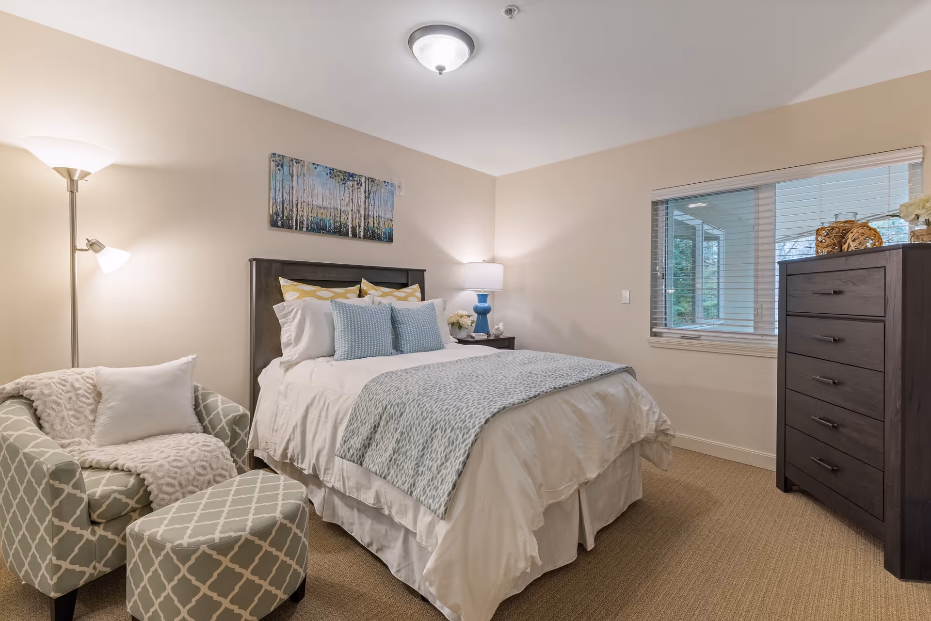A cozy bedroom with a neatly made bed featuring white and light blue bedding, a patterned armchair with a matching ottoman, a floor lamp, a nightstand with a blue lamp, and a dark wooden dresser. A window with blinds shows an outdoor view, and a painting of trees hangs above the bed.