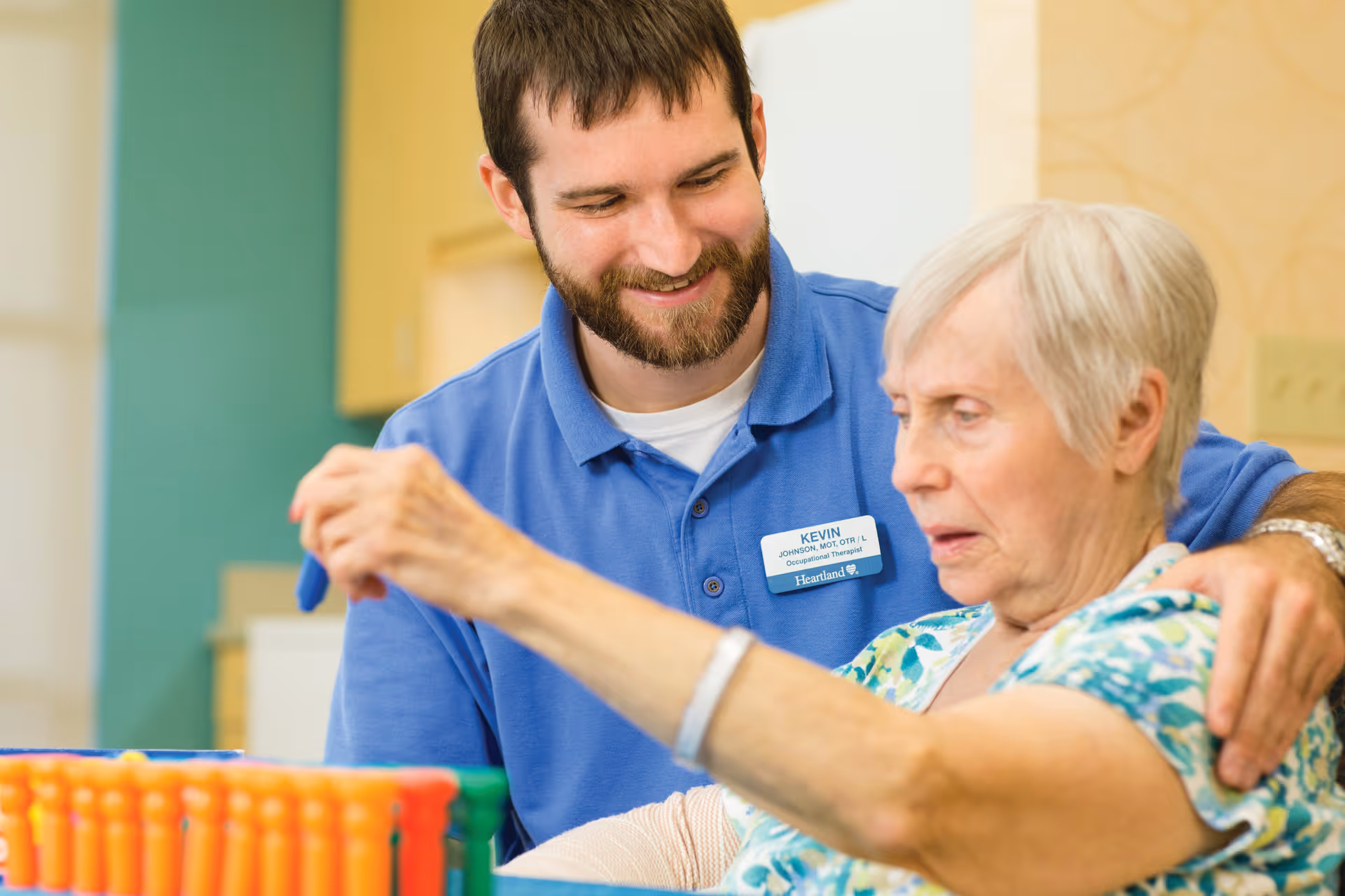 A male occupational therapist wearing a blue shirt with a name tag labeled Kevin is assisting an elderly woman with a therapeutic activity involving colorful pegs. They are indoors in a well-lit room with a green and yellow background.