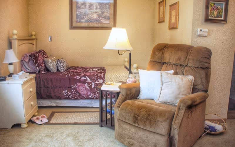 A cozy bedroom featuring a single bed with a purple floral bedspread, a white nightstand with a lamp and tissues, a beige carpet, a brown recliner chair with two pillows, a side table with a lamp, and framed artwork on the walls.