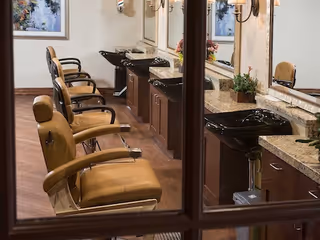 Interior view of a salon area with multiple brown leather styling chairs lined up in front of granite countertops and black sinks. Large mirrors and wall-mounted light fixtures are visible above the counters. A framed picture hangs on the wall in the background.