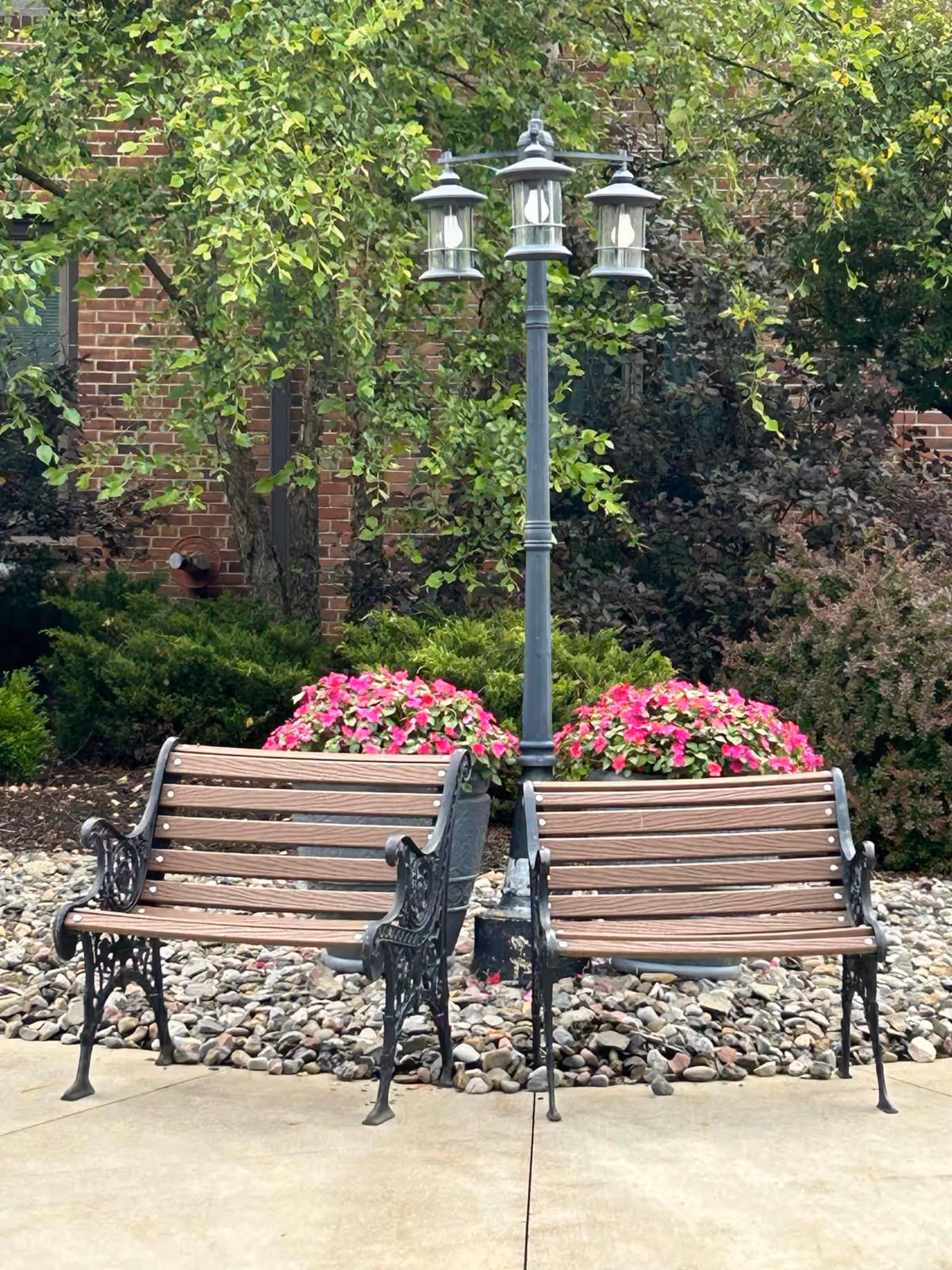 Two wooden benches with black ornate metal armrests and legs are placed on a paved area in front of a garden bed with rocks, pink flowers, green bushes, and trees. A black metal lamp post with three lanterns stands between the benches. A brick building is visible in the background.