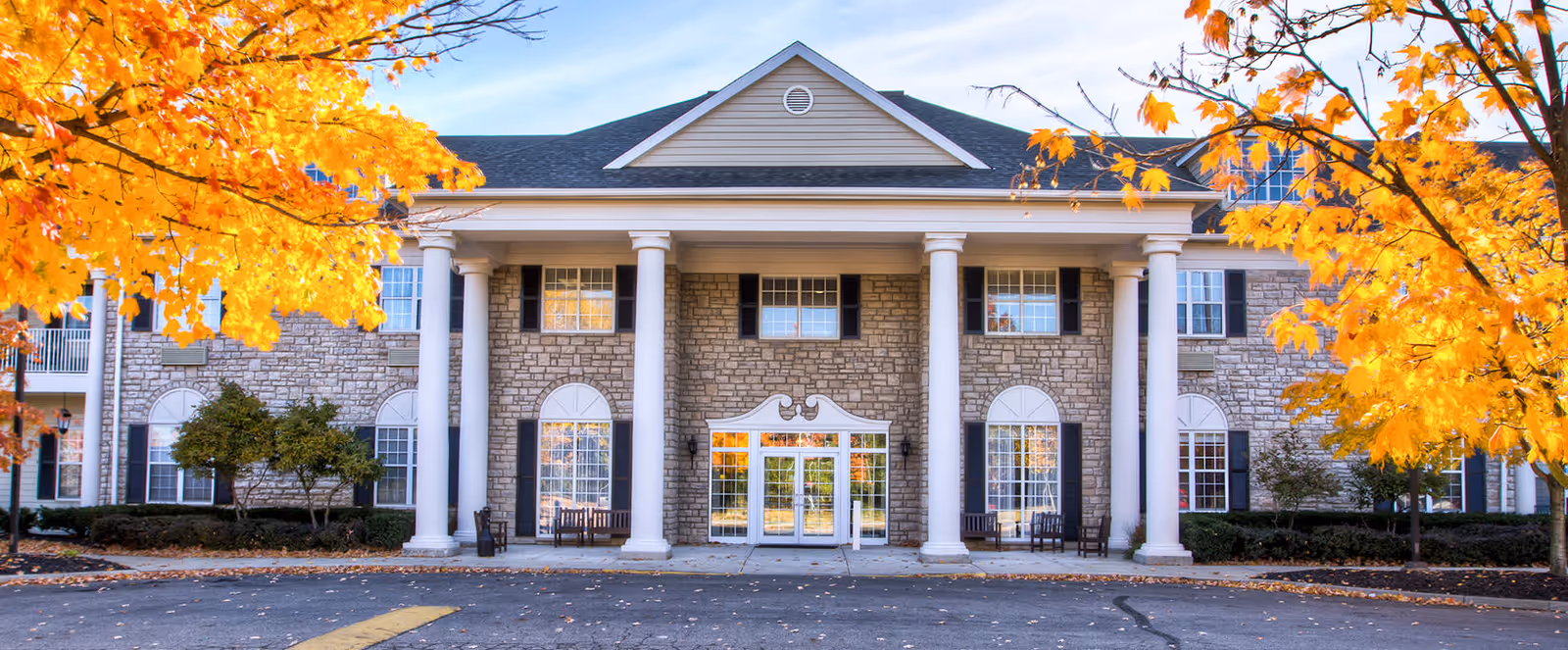 Front exterior view of a large senior living facility building with stone facade, white columns, and large windows. The scene is framed by trees with bright yellow autumn leaves.