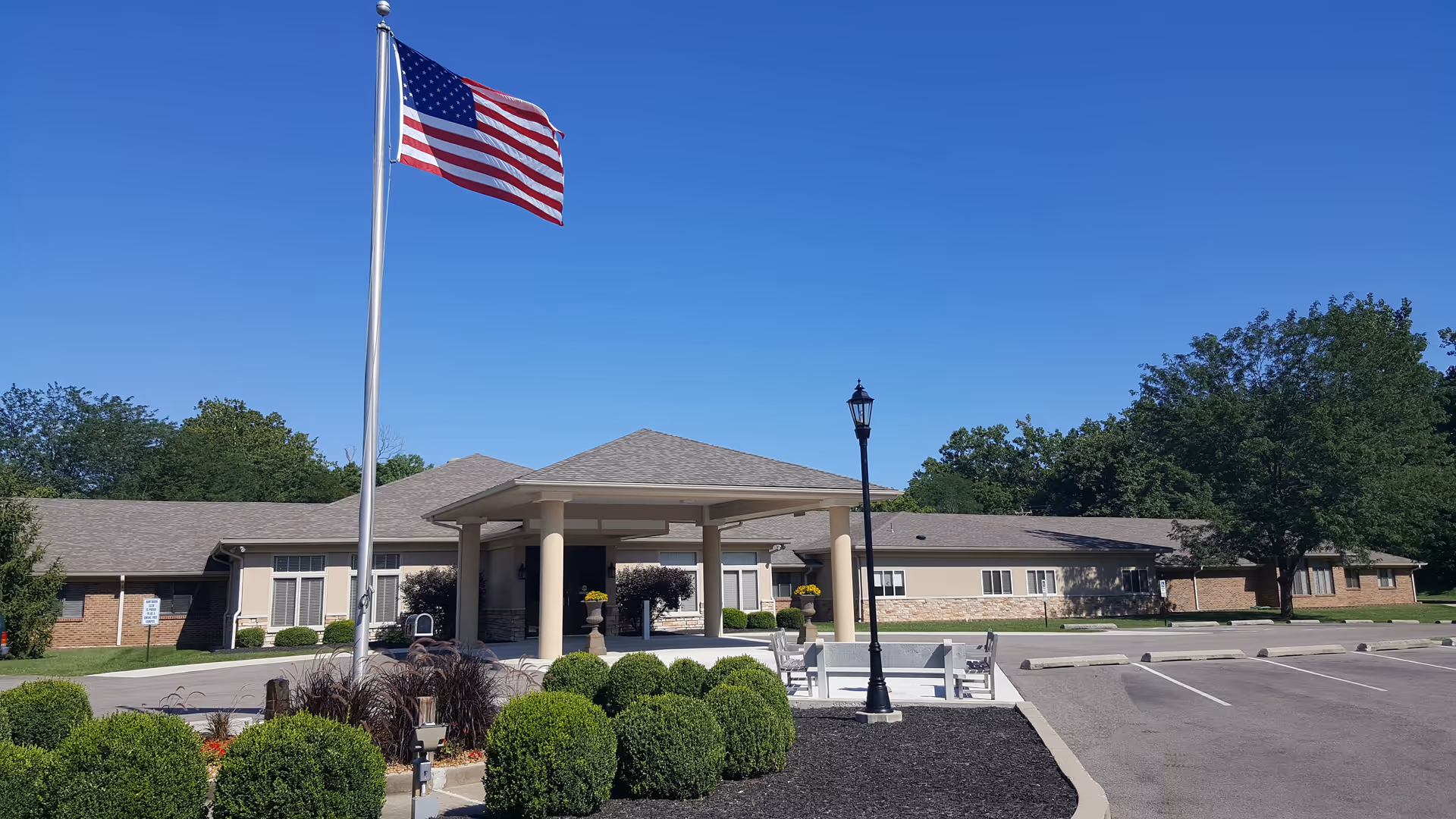 Exterior view of Hawthorn Glen Senior Living Campus building with a covered entrance, an American flag on a flagpole, neatly trimmed bushes, a lamp post, and a clear blue sky.