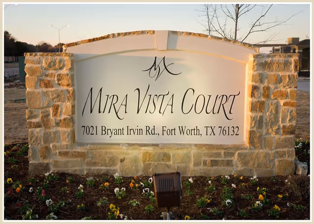 Stone monument sign for Mira Vista Court with the address 7021 Bryant Irvin Rd., Fort Worth, TX 76132, surrounded by a flower bed with small colorful flowers.