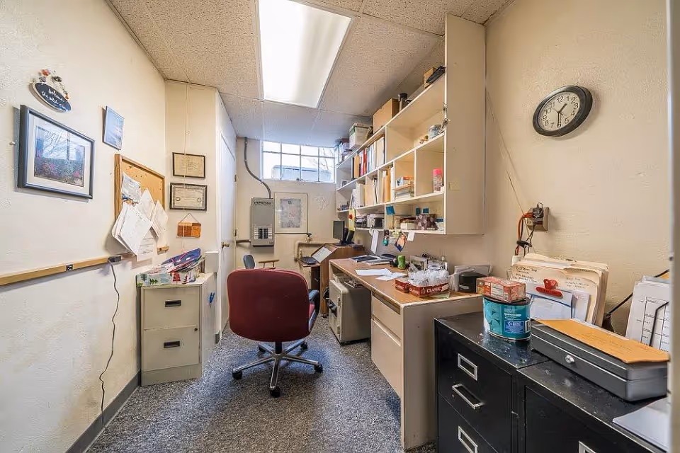 A small office room with a maroon swivel chair in front of a desk cluttered with papers, office supplies, and shelves filled with books and files. There is a filing cabinet on the left side and a clock on the wall showing the time as 12:10. The room has a window near the ceiling letting in natural light.