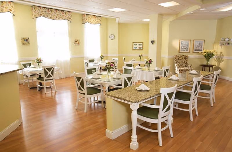 A bright and inviting dining room with several tables set with white tablecloths, napkins, and small flower arrangements. The room has light yellow walls, large windows with floral valances, wooden flooring, and white chairs with green cushions. There is a long granite countertop with chairs along one side and decorative artwork and flowers on the walls and tables.