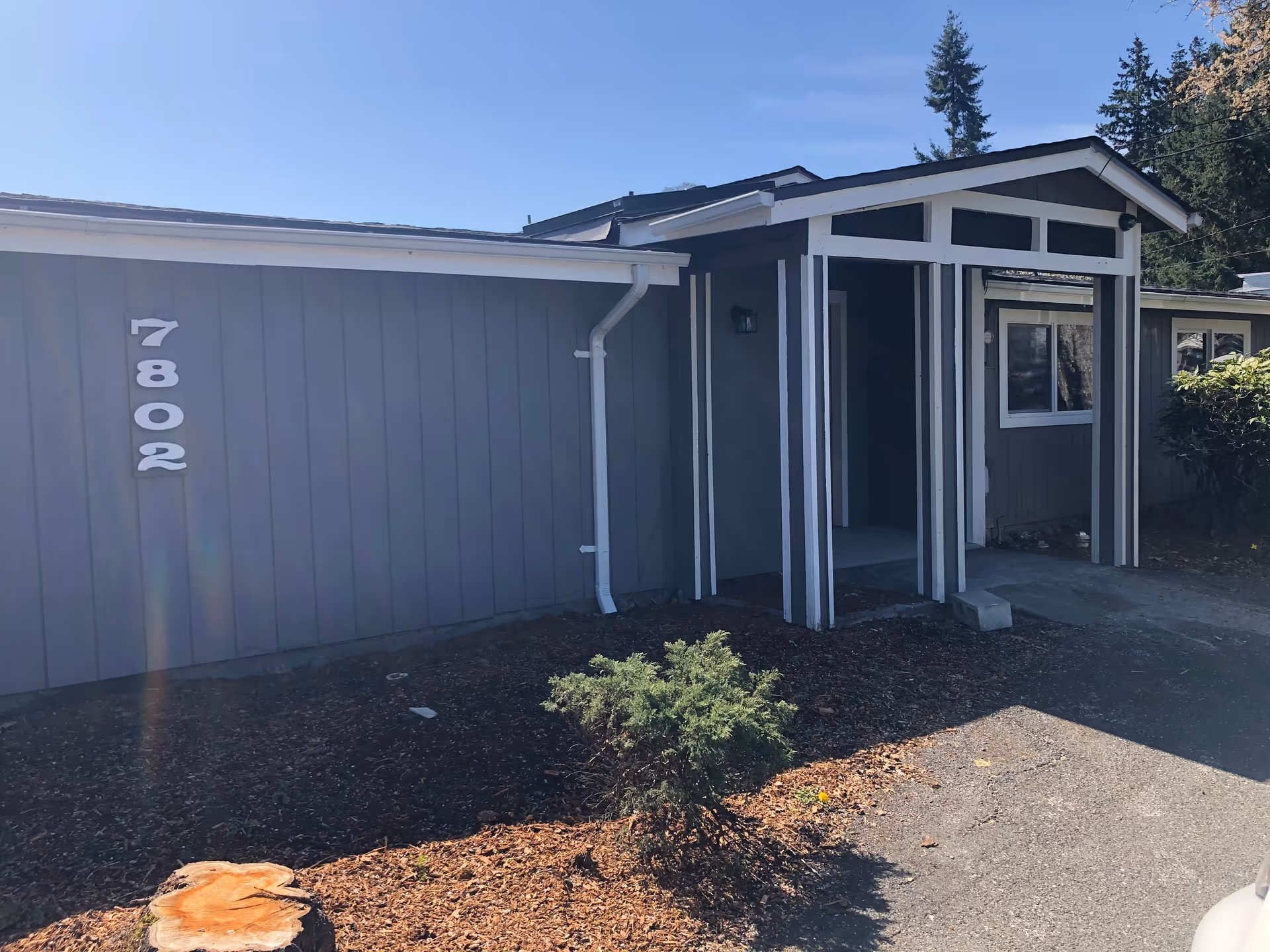 Exterior view of a single-story building with gray siding and white trim, featuring a covered entrance supported by columns. The building number 7802 is displayed on the left side of the entrance. There are some small shrubs and mulch in front of the building, and tall trees are visible in the background under a clear blue sky.