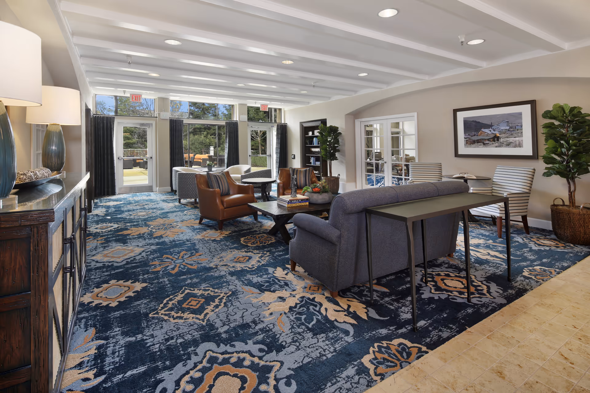 A spacious and well-lit living room area with patterned blue and beige carpet, a variety of seating including a blue sofa, brown leather armchairs, and striped chairs. There are large windows and glass doors leading to an outdoor patio area. The room features a wooden sideboard with lamps, a coffee table with books and a plant, a framed picture on the wall, and potted plants adding greenery.