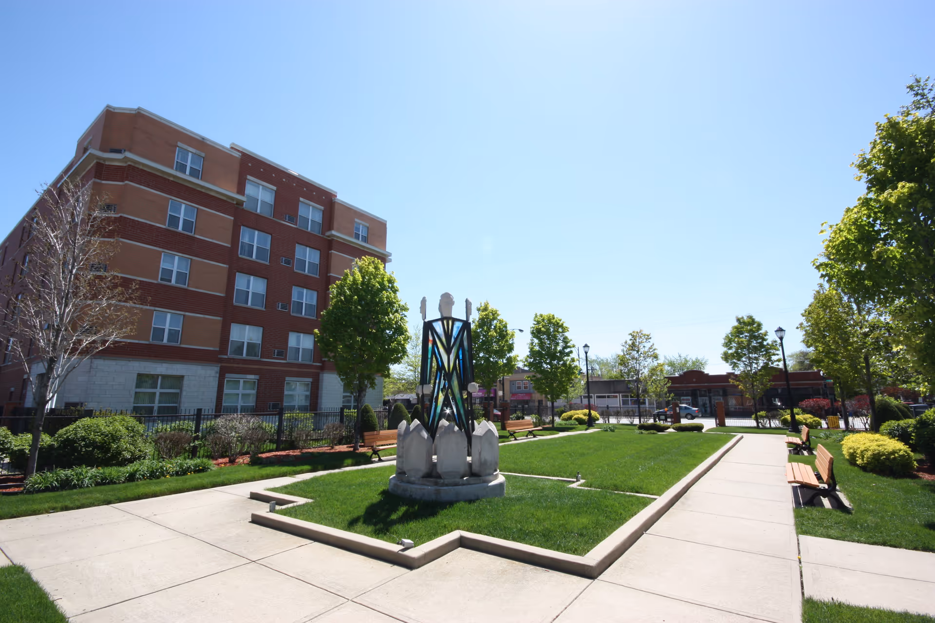Outdoor courtyard area of Churchview Supportive Living featuring a central decorative sculpture surrounded by green grass, benches along the paved walkways, trees, and a multi-story brick building in the background under a clear blue sky.