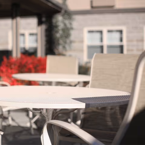 Outdoor patio area with round white metal tables and beige cushioned chairs, with a building and red foliage in the background.