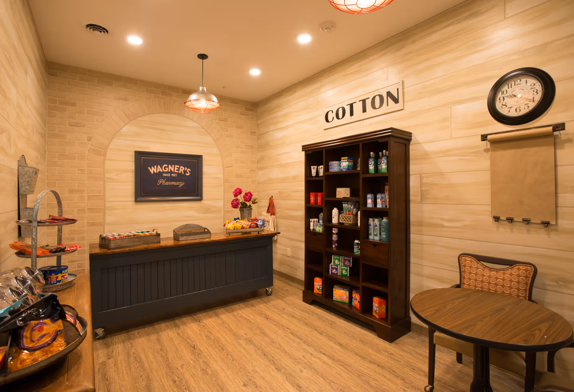 Interior view of a small pharmacy area with a wooden floor and beige walls. There is a dark wooden counter with snacks and a sign above it that reads 'WAGNER'S Pharmacy'. To the right, a tall dark wooden shelf labeled 'COTTON' holds various personal care products. A round wooden table with a patterned chair is positioned near a wall clock and a paper roll dispenser.